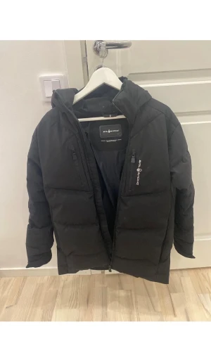 Svart pufferjacka från Sail Racing - Svart pufferjacka från Sail Racing med huva och två bröstfickor med dragkedja. Jackan har Sail Racing-logga på bröstet och patch på ärmen med texten 'Sail Antarctica'. Perfekt för kalla dagar, snygg och stilren design med quiltade sektioner.