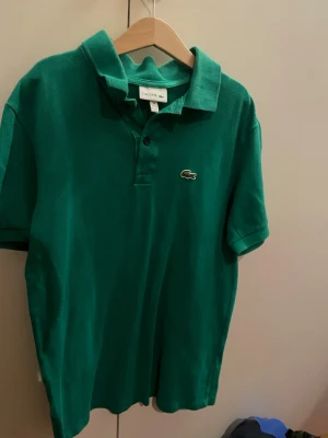 Grön pikétröja Lacoste 14 - Säljer en grön pikétröja från Lacoste med klassisk krage och korta ärmar. Tröjan har två knappar framtill och den ikoniska krokodilloggan broderad på bröstet. Materialet är mjuk bomull.