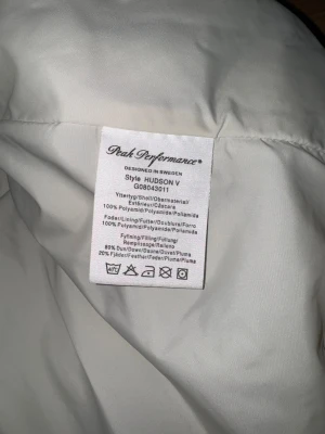 Blå dunväst Peak Performance  - Snygg blå dunväst från Peak Performance i modell Hudson V. Västen har vita paneler med röda detaljer upptill, hög krage och tryckknappar framtill. Tillverkad i polyamid med fyllning av dun och fjäder. Perfekt för lager-på-lager under kyliga dagar.