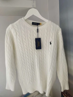 Vit kabelstickad tröja Polo Ralph Lauren - Snygg vit kabelstickad tröja från Polo Ralph Lauren med klassisk rund hals och broderad logga på bröstet. Tröjan har långa ärmar och ribbade muddar vid ärmslut och nederkant. Perfekt för dig som gillar stilrena och tidlösa plagg.