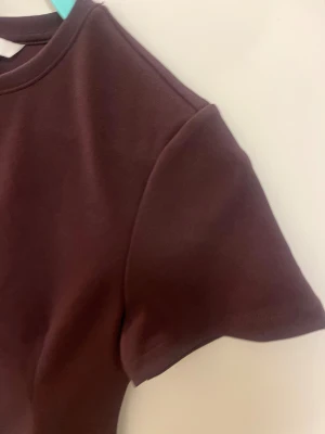 Vinröd kortärmad topp från H&M - Säljer en vinröd topp från H&M med korta ärmar och rund halsringning. Toppen har en figurnära passform och är tillverkad i ett mjukt, stretchigt material som känns skönt mot huden. Perfekt att styla med jeans eller kjol för en clean look.