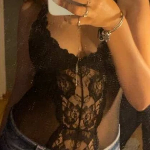 Svart spetsig body med blommönster - Säljer en svart body med blommig spets och djup v-ringning. Toppen är tight och har tunna axelband, transparent mesh på sidorna och detaljerad spets framtill. Perfekt för dig som vill ha en edgy och snygg look.