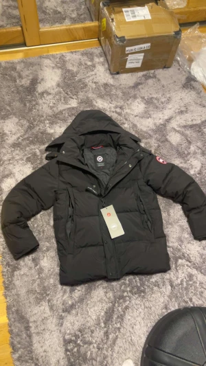Canada Goose Wyndham  - Svart dunjacka från Canada Goose med huva och klassisk logga på ärmen. Jackan har dragkedja, knappar och två stora fickor framtill. Puffad design som ger en schysst siluett och håller dig varm under kalla dagar. Perfekt för dig som gillar stil och funktion.