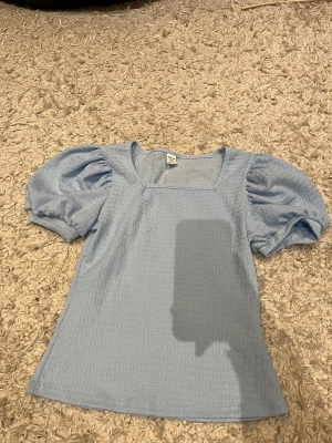 Ljusblå topp med puffärm från SHEIN - Supersöt ljusblå topp från SHEIN med puffiga korta ärmar och fyrkantig ringning. Toppen har en lätt strukturerad yta och är i ett mjukt, stretchigt material. Perfekt för dig som gillar en feminin och trendig stil.