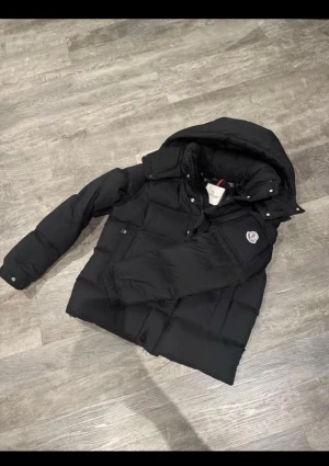 Svart Moncler pufferjacka med huva - Säljer en svart pufferjacka från Moncler med stor huva och snygga detaljer som logotyp på ärmen. Jackan har dragkedja, knappar och flera fickor framtill. Perfekt för kalla dagar och har en riktigt schysst, puffig look.