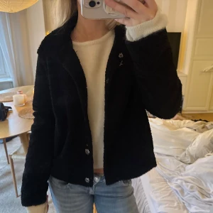 Svart teddyjacka med knappar - Mysig svart teddyjacka med krage och knäppning framtill. Jackan har en kortare, normal passform och är tillverkad i fluffigt teddy-material / pile jacka. Från Nelly strl s