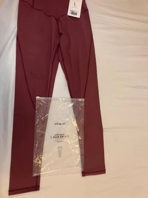 Vinröda leggings från La Nikar, stl L - Snygga vinröda leggings från La Nikar i storlek L. De har hög midja och är tillverkade i ett stretchigt syntetmaterial som sitter tight och bekvämt. Perfekta för träning eller chill, med coola sömdetaljer längs benen för extra stil.