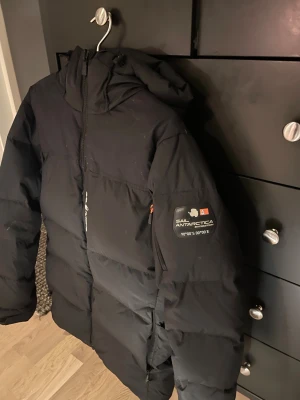 Svart dunjacka Sail Racing - Svart dunjacka från Sail Racing med huva och snygg expedition-patch på ärmen. Jackan är använd 1 vinter då ja växt ut den, inte en ända skada/slitning på den. Kostar 4500kr ny
