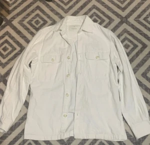Vit overshirt med knappar - Säljer en stilren vit overshirt från Arket i bomull. Skjortan har klassisk krage, två bröstfickor med lock och knappar framtill. Perfekt att bära som lager över t-shirt eller tröja. Långärmad och med en clean look.