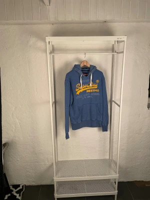 Blå Superdry hoodie med tryck - Blå hoodie från Superdry med stort gult och vitt tryck på bröstet. Tröjan har huva med vita snören, ribbade muddar och en klassisk känguruficka framtill. Perfekt för dig som gillar streetwear och snygga loggor.