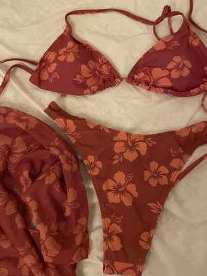 Blommig bikini med snörning - Säljer en snygg bikini med stora orangea hibiskusblommor. Bikinin har trekantsformad topp med snörning och matchande trosa med högt skurna ben. Kommer med en duk till midjan. Aldrig använt och säljs i befintlig skick