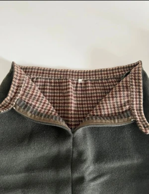 Mörk olivgrön ullkjol med rutiga detaljer - Mörk olivgrön långkjol i ull med rutigt mönster i beige, brun och vinröd längs nederkanten och insidan av midjan. Kjolen har en klassisk rak passform, knapp och dragkedja i midjan samt diskreta fickor i sidorna. Köpt från butik i Italien. Helt ny utan prislapp.