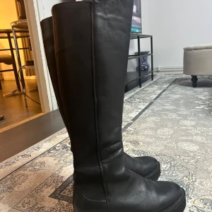 Svarta höga stövlar i äkta skinn - Säljer ett par svarta höga stövlar med grov sula och rund tå. Stövlarna är tillverkade i äkta skinn och har elastiska paneler längs skaftet för extra komfort. Perfekta för dig som gillar en stilren look med edge. Snygga till både jeans och kjol. köptes för 1800:- säljer dessa för 500:-