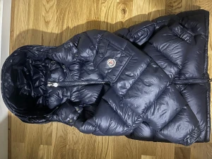 Mörkblå Moncler dunjacka med huva - Säljer en mörkblå dunjacka från Moncler med stor huva och bröstficka med logotyp. Jackan har två sidofickor med dragkedja och brunt foder med serietidningsdetalj. Perfekt för kalla dagar och har en riktigt puffad look. Kvitto finns.