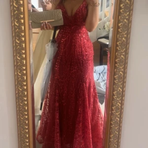 Röd glittrig balklänning - Säljer en röd långklänning med glittriga paljetter, djup v-ringning och smala axelband. Klänningen har öppen rygg med snygg snörning och figurnära passform upptill som faller ut i ett vackert svepande tyg. Perfekt för bal💓Storlek 38, jag är runt 167 och har klackar i bilderna💓tryck gärna på köp nu