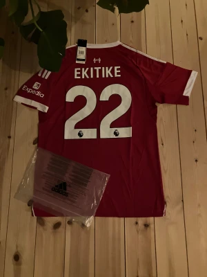 Liverpool röd fotbollströja Adidas - Liverpool FC matchtröja från Adidas i rött med vita detaljer och tryck. Kortärmad med klubbmärke, sponsorloggor och spelarnamn Ekitike samt nummer 22 på ryggen. Tillverkad i lätt och ventilerande material, perfekt för fotboll eller häng med polarna.