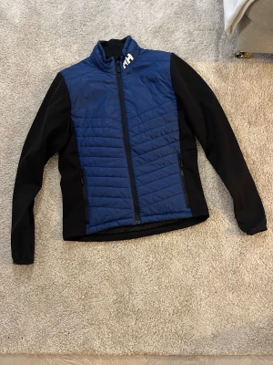 Blå och svart pufferjacka Helly Hansen - Snygg pufferjacka från Helly Hansen i blått och svart med HH-logga på bröstet och ryggen. Jackan har quiltad front, dragkedja och två sidofickor med zip. Perfekt för kyliga dagar och har en sportig look med hög krage. Den håller dog varm även vid minus grader med en hoodie under.