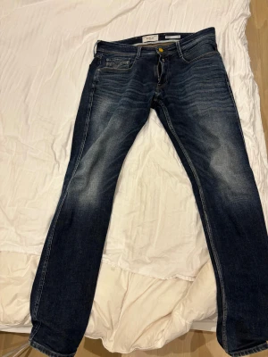 Replay Anbass slim fit jeans blå - Säljer ett par mörkblå Replay Anbass slim fit jeans med snygga slitningar och klassisk femficksdesign. Jeansen har gul knapp, kontrastsömmar och är tillverkade i stretchig denim för en bekväm passform. Perfekta för dig som gillar en modern och stilren look.