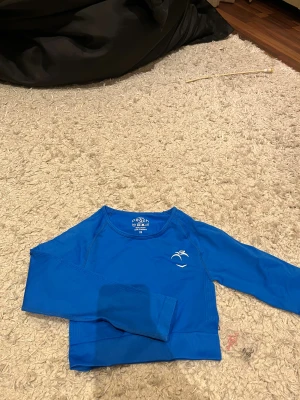 Blå långärmad träningströja Peach XS - Snygg blå långärmad träningströja från Peach i storlek XS. Tröjan är croppad och har ett stilrent tryck på bröstet. Tillverkad i mjukt och stretchigt material med 90% nylon och 10% elastan, perfekt för gymmet eller sportaktiviteter.
