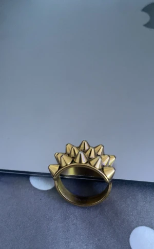 Guldfärgad ring med nitar från Edblad - Cool och chunky guldfärgad ring från Edblad med flera konformade nitar som ger en edgy vibe. Ringen är bred och har en unik design som sticker ut. Perfekt för dig som gillar statement-smycken och vill ha något som syns.