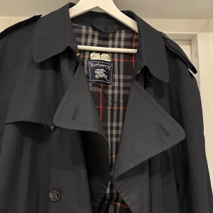 Svart trenchcoat från Burberrys - Svart trenchcoat från Burberry, modell kensington, med klassiskt rutigt foder i grått, vitt och rött. Dubbelknäppning, bälte i midjan med metallspänne och justerbara remmar vid ärmslut. Måtten är följande:   Bredd (över bröstet): 52 cm   Axelbredd: 45 cm   Längd: 122 cm   Ärmlängd: 65 cm   Armbredd: 13,5 cm