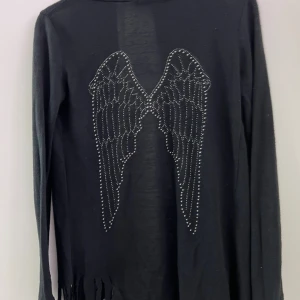 Svart långärmad kofta med vingar - Svart långärmad kofta med coola vingmotiv i silverfärgade nitar på ryggen. Koftan har en öppen front och fransar nedtill som ger en edgy vibe. Perfekt för dig som gillar att sticka ut med detaljer och vill ha något extra till din outfit.