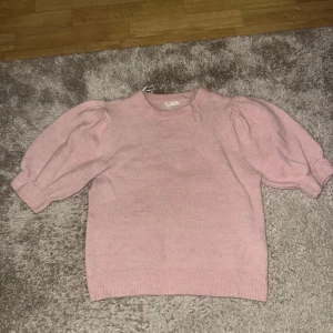 Ljusrosa stickad tröja Gina Tricot - Supersöt ljusrosa stickad tröja från Gina Tricot med puffiga korta ärmar och ribbade muddar. Tröjan har rund hals och är i mjukt material som känns skönt mot huden. Perfekt för dig som gillar en feminin och trendig look.