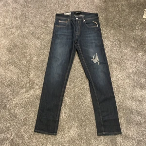 Replay Grover jeans mörkblå herr - Snygga Replay Grover jeans i mörkblå tvätt med klassisk femficksdesign och slitning på höger lår. Jeansen har raka ben och kontrastsömmar, samt Replay-logga på linningen. Perfekt för dig som gillar en cool och avslappnad stil.