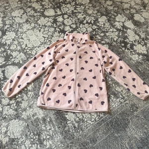 Rosa fleecejacka med hjärtan från H&M - Supergullig rosa fleecejacka från H&M med mörklila hjärtmönster. Jackan har dragkedja framtill, hög krage och långa ärmar. Perfekt för kyliga dagar när du vill vara både varm och söt.