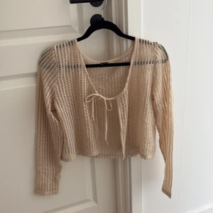 Beige mesh blus från Vero Moda - Säljer en beige, transparent meshblus från Vero Moda i storlek M. Blusen har lång ärm, croppad passform och en söt knytning framtill vid halsen. Perfekt för lager-på-lager och har en luftig, modern look med snyggt rutmönstrad struktur.