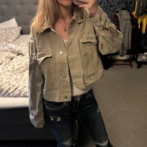 Olivgrön croppad jeansjacka från Zara - Snygg croppad jeansjacka från Zara Basic i olivgrön denim. Jackan har stora bröstfickor med lock, klassisk krage och knäppning framtill. Perfekt för dig som gillar en avslappnad och trendig stil.