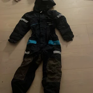 Marinblå overall från Didriksons stl 130 - Marinblå overall från Didriksons med blå detaljer och reflexer. Overallen har huva, dragkedja framtill och förstärkta partier på knän och rumpa. Perfekt för kalla dagar med sitt vattenavvisande material och praktiska fickor med tryckknappar. 