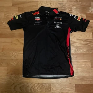 Red Bull Racing svart skjorta L - Svart Red Bull Racing skjorta med tryckta loggor från Aston Martin, Honda, Mobil 1 och Puma. Skjortan har krage, korta ärmar och röda detaljer på sidan. Tillverkad i 100% polyester, perfekt för dig som gillar motorsport och snygga teamkläder.