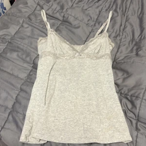 Grå linnetopp med spetsdetaljer - Säljer en ljusgrå linnetopp med tunna axelband och fina spetsdetaljer längs urringningen. Toppen har en mjuk och följsam passform