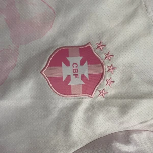 Vit och rosa Neymar CBF fotbollströja - Snygg fotbollströja från Nike med CBF-emblem och Neymar 10 på ryggen. Tröjan är vit med rosa detaljer och har ett stort tryck av Kristusstatyn och Rio de Janeiro på framsidan. Tillverkad i lätt Dri-FIT material som andas, perfekt för träning eller match.