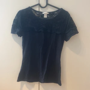 Svart spetsblus från H&M - Säljer en svart blus från H&M i storlek S. Toppen har korta ärmar och en snygg spetsdetalj över axlar och bröst, vilket ger en cool och feminin vibe. Perfekt att styla med jeans eller kjol för en trendig look.