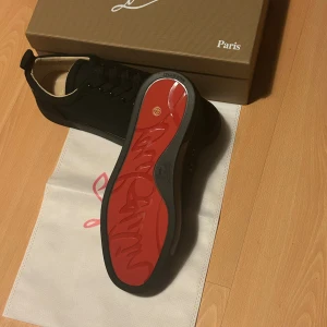 Säljer ett till par Svarta sneakers från Christian Louboutin - Snygga svarta sneakers från Christian Louboutin med klassisk röd signatursula och diskret guldfärgad logga på innersulan. Skorna har rund tå, snörning och är tillverkade i skinn med en stilren, minimalistisk look. Perfekta för dig som gillar exklusiva detaljer.