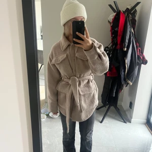 Beige kappa med bälte från NA-KD - Säljer en beige kappa med knappar och bälte i midjan. Jackan har stora fickor framtill. Pris kan diskuteras!