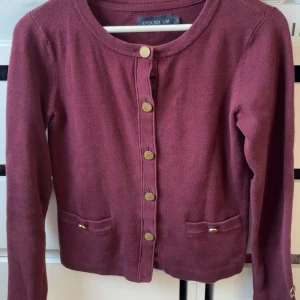 Vinröd kofta från Stockh LM - Vinröd kofta med rund hals och guldknappar framtill från Stockh LM. Koftan har två fickor med dekorativa knappar och är tillverkad i ett mjukt stickat material. Perfekt för lager-på-lager och har långärmad passform.