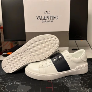 Valentino Garavani Open - Snygga Valentino Garavani Open sneakers i vitt skinn med bred svart läderpanel över snörningen och coola nitar på sidan av sulan. Platt sula med mönstrad undersida och rund tå. Logotyp i guld på plösen och innersulan. Helt nya och oandvända! Stl 38. Perfekt för dig som gillar exklusiv streetstyle.