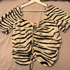 Zebramönstrad snörad blus - Säljer en snygg zebramönstrad blus med svartvita ränder och puffiga korta ärmar. Toppen har snörning framtill som ger en cool, rynkad effekt. Från HM, storlek XL men passa L också. Dragkedja på sidan och du kan spänna snörningen som du vill. Oanvänd 