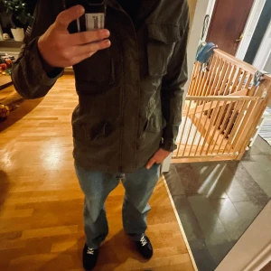 Olivgrön army parkajacka med patchar - Säljer en olivgrön parkajacka i bomull med coola militärpatchar, amerikansk flagga på ärmen och stora tryck med 'ARMY' på ryggen. Jackan har fyra fickor framtill, hög krage med spänne och klassisk army-vibe. Perfekt för dig som gillar utility-stil.