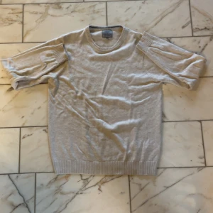 Beige/vit tröja från Morley - Snygg beige stickad tröja från Morley med korta ärmar och ribbade muddar. Tröjan har rund hals och ett diskret broderat emblem på bröstet. Perfekt för dig som gillar stilrena och klassiska plagg med en modern touch.