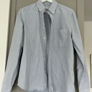 Randig blåvit skjorta från ASKET - Säljer en klassisk blåvit randig skjorta från ASKET. Skjortan har lång ärm, knappar framtill och en bröstficka. Tillverkad i bomull och har en tidlös look med smala vertikala ränder. Perfekt för dig som gillar stilrena plagg med enkel design.