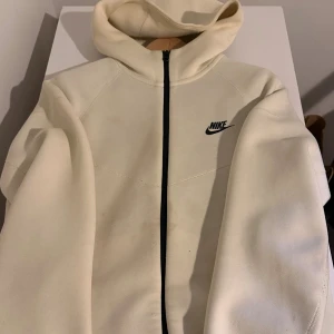 Beige hoodiejacka från Nike, stl S - Snygg beige hoodiejacka från Nike med svart logga på bröstet och hel dragkedja framtill. Jackan har huva och är tillverkad i mjukt material, perfekt för en sportig och avslappnad stil.