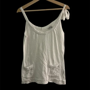 Vit linnetopp med fickor och knyt - Säljer en söt vit linnetopp från Morgan  med smala axelband, varav det ena har en knyt-detalj. Toppen har två små fickor framtill med volangkant och dekorativt band. 