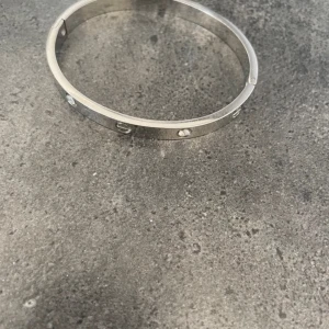 armband i silver - Snyggt och stilrent armband i silverfärgad metall med ikoniska skruvdetaljer och infattade stenar runt om. Armbandet har en rund, klassisk form och är perfekt för dig som gillar exklusiva accessoarer med lyxig känsla.