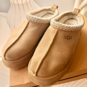 Beige UGG tazz slip-ons med plattform - Säljer ett par beige slip-on skor från UGG med plattform. Skorna har mocka på utsidan, mjuk päls på insidan och dekorativt mönster runt öppningen.  Sparsamt använda. Tyvärr blev dom för stora storlek 39