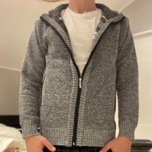 Cardigan från Montéva  - Säljer en grå stickad hoodie med huva och hel dragkedja framtill. Tröjan har ett diskret mönster, ribbade muddar och är långärmad. Perfekt för kyliga dagar och har en avslappnad passform.