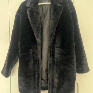 Svart pälsjacka från rut&circle - Säljer en svart pälsjacka från rut&circle med klassisk krage och långärmad design. Jackan är oversized och har ett mjukt, fluffigt material som ger en lyxig vibe. Perfekt för kalla dagar när du vill vara både varm och snygg.